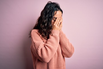 Young beautiful woman with curly hair wearing casual sweater over isolated pink background with sad expression covering face with hands while crying. Depression concept.