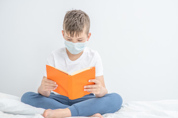 Smart boy wearing medical mask sits on a bed and reads a book. Quarantine and coronavirus epidemic concept. Empty space for text