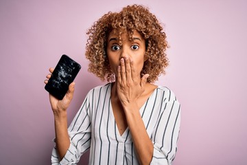 Young african american curly woman holding broken smartphone showing craked screen cover mouth with...