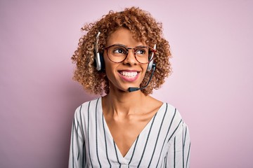 African american curly call center agent woman working using headset over pink background looking...
