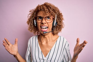 African american curly call center agent woman working using headset over pink background crazy and mad shouting and yelling with aggressive expression and arms raised. Frustration concept.