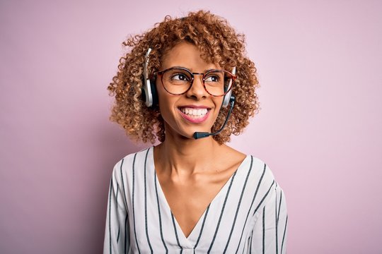 African american curly call center agent woman working using headset over pink background looking away to side with smile on face, natural expression. Laughing confident.