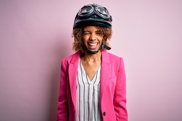 African american motorcyclist woman with curly hair wearing moto helmet over pink background winking looking at the camera with sexy expression, cheerful and happy face.