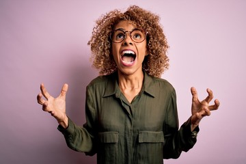 Young beautiful african american woman wearing casual shirt and glasses over pink background crazy and mad shouting and yelling with aggressive expression and arms raised. Frustration concept.