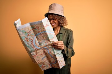 African american tourist woman wearing explorer hat and water canteen holding city map with a happy face standing and smiling with a confident smile showing teeth
