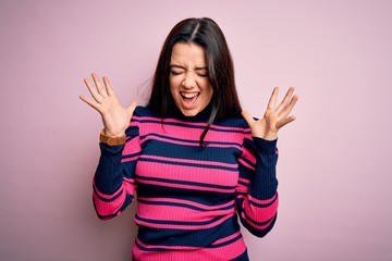 Young brunette elegant woman wearing striped shirt over pink isolated background celebrating mad and crazy for success with arms raised and closed eyes screaming excited. Winner concept