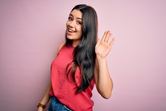 Young brunette woman wearing casual summer shirt over pink isolated background Waiving saying hello happy and smiling, friendly welcome gesture