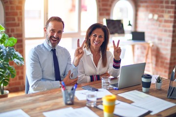 Middle age beautiful business workers working together using laptop at the office smiling with happy face winking at the camera doing victory sign. Number two.