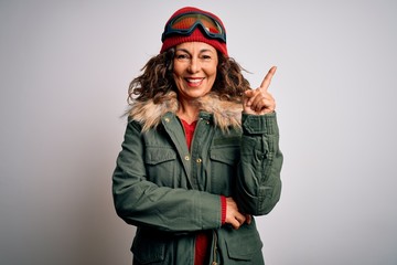 Middle age skier woman wearing snow sportswear and ski goggles over white background with a big smile on face, pointing with hand and finger to the side looking at the camera.