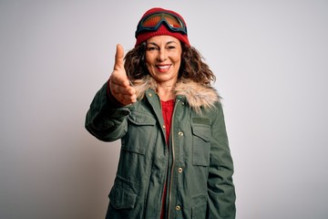 Middle age skier woman wearing snow sportswear and ski goggles over white background smiling friendly offering handshake as greeting and welcoming. Successful business.
