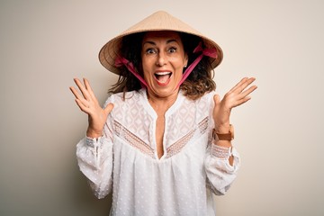 Middle age brunette woman wearing asian traditional conical hat over white background celebrating crazy and amazed for success with arms raised and open eyes screaming excited. Winner concept