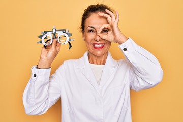 Middle age senior oculist woman holding optometrical glasses for vision correction with happy face smiling doing ok sign with hand on eye looking through fingers