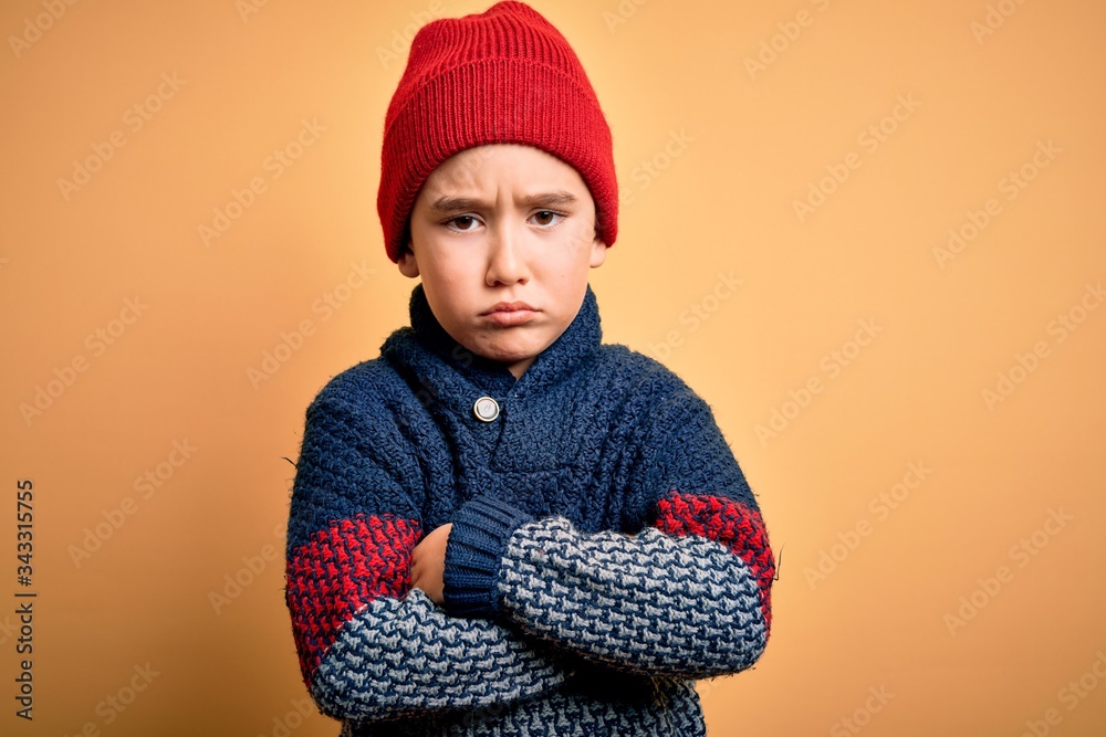 Poster Young little boy kid wearing wool cap and winter sweater over yellow isolated background skeptic and nervous, disapproving expression on face with crossed arms. Negative person.