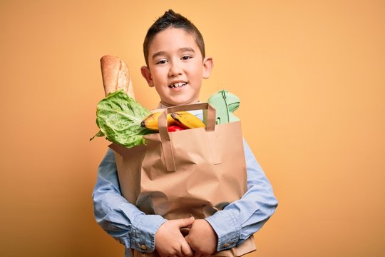 Adorable toddler holding paper bag with food standing over isolated yellow background