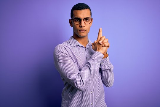 Handsome african american man wearing striped shirt and glasses over purple background Holding symbolic gun with hand gesture, playing killing shooting weapons, angry face