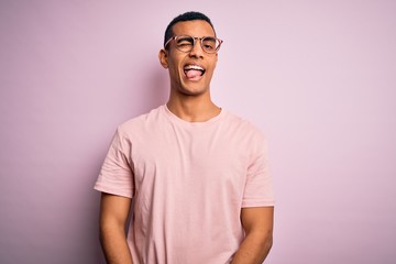 Obraz premium Przystojny Afroamerykanin ubrany na co dzień t-shirt i okulary na różowym tle mrugając patrząc w kamerę z seksowną ekspresją, wesołą i szczęśliwą twarzą.