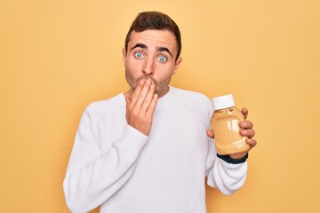 Young handsome man holding bottle of mustard sauce condiment over yellow background cover mouth...
