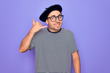 Handsome man with blue eyes wearing striped t-shirt and french beret over purple background smiling doing phone gesture with hand and fingers like talking on the telephone. Communicating concepts.