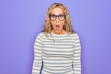 Naklejka premium Beautiful blonde woman wearing casual striped t-shirt and glasses over purple background afraid and shocked with surprise and amazed expression, fear and excited face.