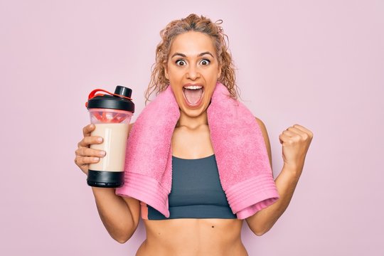 Beautiful blonde sporty woman doing sport wearing towel holding shaker with protein beverage screaming proud, celebrating victory and success very excited with raised arms