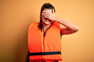 Young beautiful brunette woman wearing orange safe lifejacket over yellow background smiling and laughing with hand on face covering eyes for surprise. Blind concept.