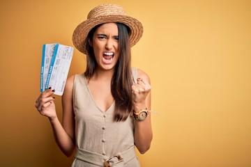 Young beautiful brunette tourist woman on vacation wearing hat holding boarding pass annoyed and frustrated shouting with anger, crazy and yelling with raised hand, anger concept