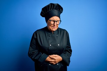 Senior beautiful grey-haired chef woman wearing cooker uniform and hat over blue background with hand on stomach because nausea, painful disease feeling unwell. Ache concept.