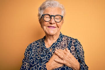 Senior beautiful grey-haired woman wearing casual shirt and glasses over yellow background smiling with hands on chest with closed eyes and grateful gesture on face. Health concept. - obrazy, fototapety, plakaty