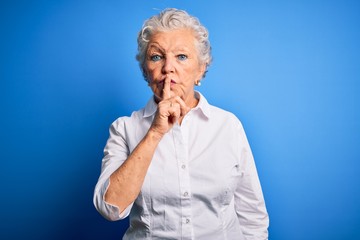 Senior beautiful woman wearing elegant shirt standing over isolated blue background asking to be quiet with finger on lips. Silence and secret concept.