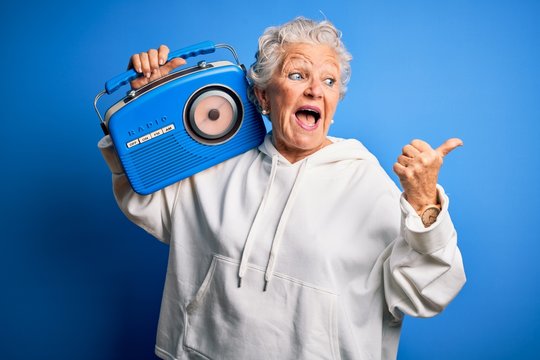 Senior beautiful woman holding vintage radio standing over isolated blue background pointing and showing with thumb up to the side with happy face smiling
