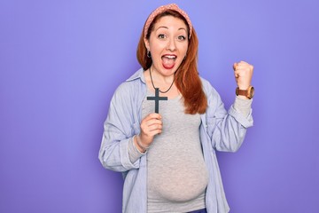 Young beautiful redhead pregnant woman expecting baby holding christian cross screaming proud and celebrating victory and success very excited, cheering emotion