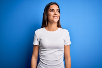Obraz premium Young beautiful brunette woman wearing white casual t-shirt standing over blue background looking away to side with smile on face, natural expression. Laughing confident.