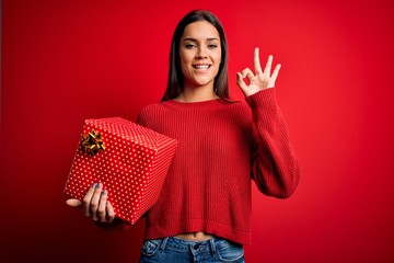 Young beautiful brunette woman holding birthday gift over isolated red background doing ok sign with fingers, excellent symbol