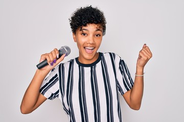 Beautiful african american afro singer woman singing using microphone over white background screaming proud and celebrating victory and success very excited, cheering emotion