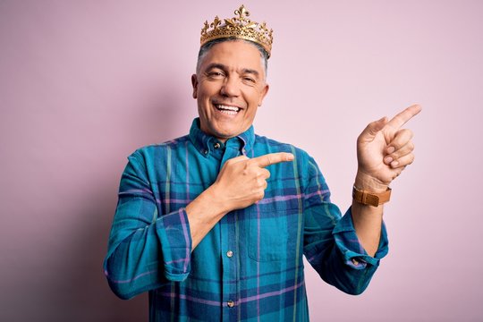 Middle age handsome grey-haired man wearing golden crown of king over pink background smiling and looking at the camera pointing with two hands and fingers to the side.