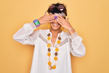 Young beautiful african american afro hippie woman wearing sunglasses and accessories covering eyes with hands smiling cheerful and funny. Blind concept.