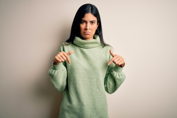 Young beautiful hispanic woman wearing green winter sweater over isolated background Pointing down looking sad and upset, indicating direction with fingers, unhappy and depressed.