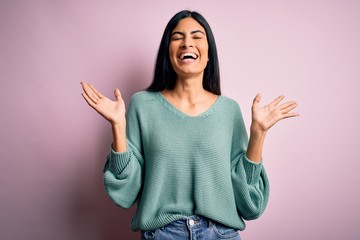 Young beautiful hispanic fashion woman wearing green sweater over pink background celebrating mad and crazy for success with arms raised and closed eyes screaming excited. Winner concept