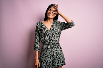 Young beautiful hispanic fashion woman wearing casual dress over pink background doing ok gesture with hand smiling, eye looking through fingers with happy face.