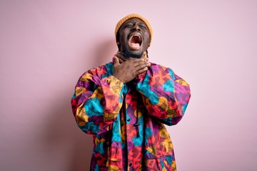 Young handsome african american man wearing colorful coat and cap over pink background shouting and suffocate because painful strangle. Health problem. Asphyxiate and suicide concept.