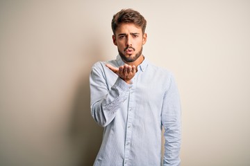 Young handsome man with beard wearing striped shirt standing over white background looking at the camera blowing a kiss with hand on air being lovely and sexy. Love expression.