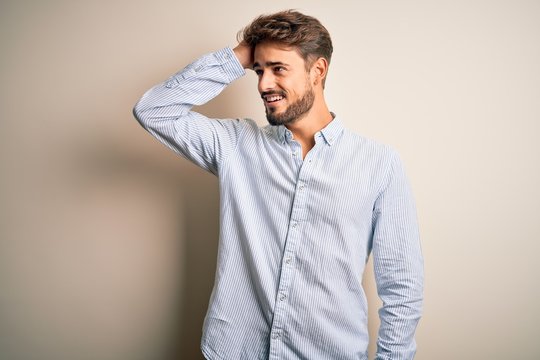 Young Handsome Man With Beard Wearing Striped Shirt Standing Over White Background Smiling Confident Touching Hair With Hand Up Gesture, Posing Attractive And Fashionable