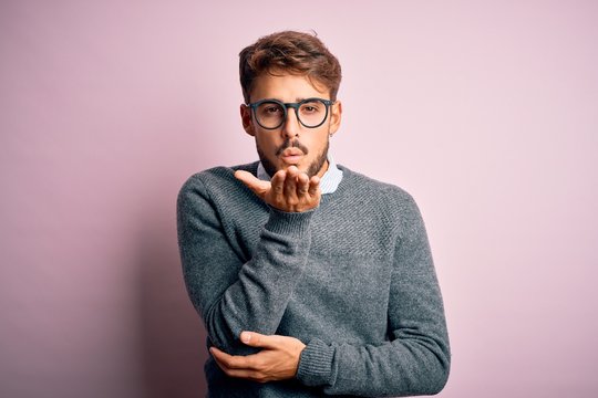 Young handsome man with beard wearing glasses and sweater standing over pink background looking at the camera blowing a kiss with hand on air being lovely and sexy. Love expression.