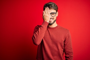 Young handsome man with beard wearing glasses and sweater standing over red background covering one eye with hand, confident smile on face and surprise emotion.