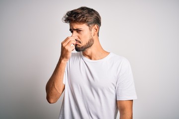 Young handsome man with beard wearing casual t-shirt standing over white background smelling something stinky and disgusting, intolerable smell, holding breath with fingers on nose. Bad smell