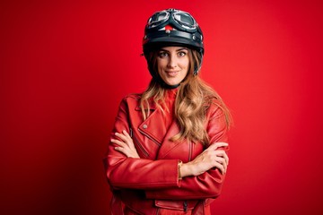 Young beautiful brunette motrocyclist woman wearing moto helmet over red background happy face smiling with crossed arms looking at the camera. Positive person.