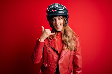 Young beautiful brunette motrocyclist woman wearing moto helmet over red background smiling doing phone gesture with hand and fingers like talking on the telephone. Communicating concepts.