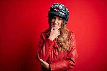 Young beautiful brunette motrocyclist woman wearing moto helmet over red background looking confident at the camera smiling with crossed arms and hand raised on chin. Thinking positive.