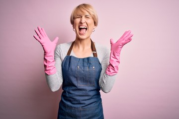 Young blonde cleaner woman with short hair wearing apron and gloves over pink background celebrating mad and crazy for success with arms raised and closed eyes screaming excited. Winner concept