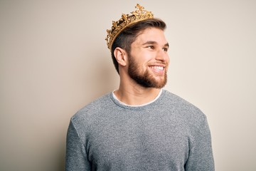 Young blond man with beard and blue eyes wearing golden crown of king looking away to side with smile on face, natural expression. Laughing confident.